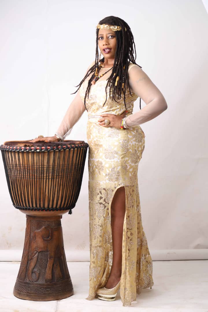 ElnanaAfrika in a gold beaded gown standing beside a large djembe drum